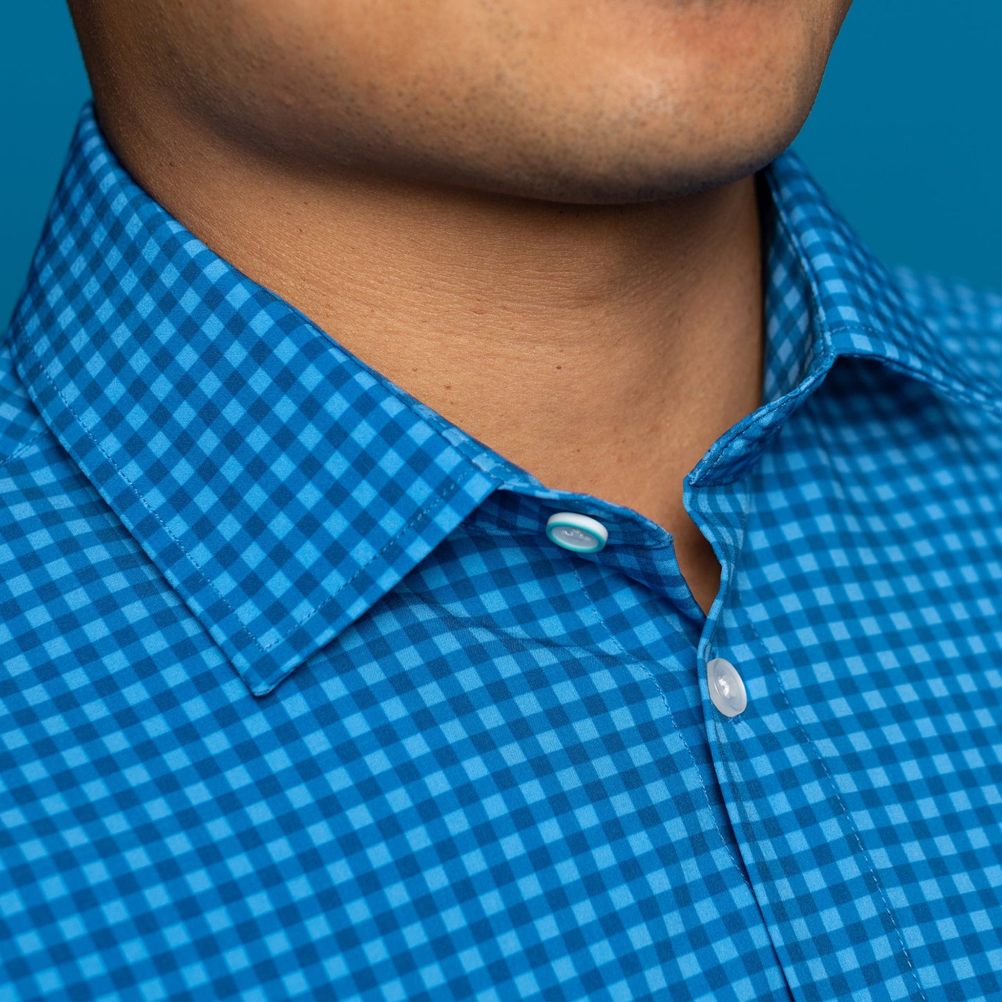 Detail of man wearing navy and royal blue gingham shirt. Top button has blue ring