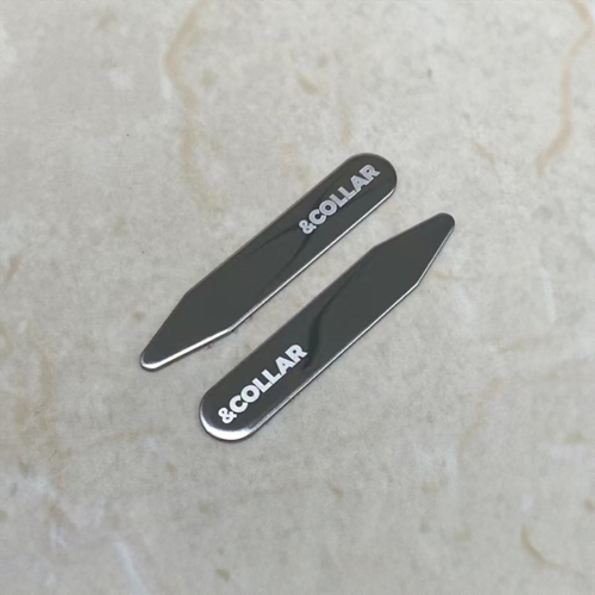 Two metallic collar stays with ' COLLAR' branding on a light marble background