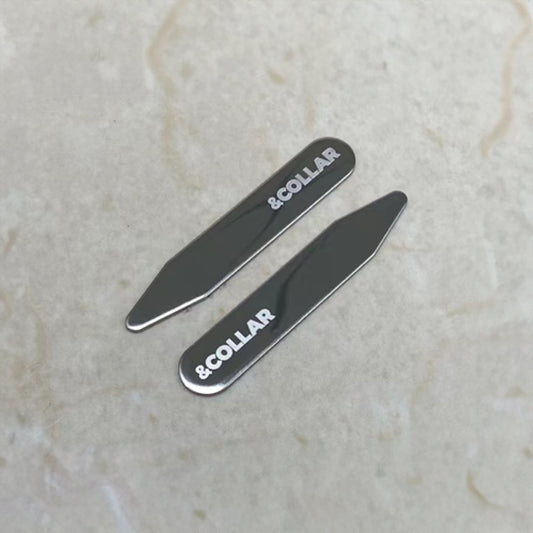 Two metallic collar stays with ' COLLAR' branding on a light marble background